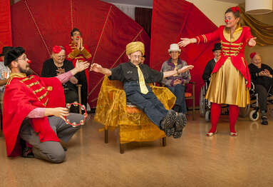 Zwei Clowns präsentieren den Auftritt eines Seniors, der auf einem Nagelbrett in einem Stuhl balanciert