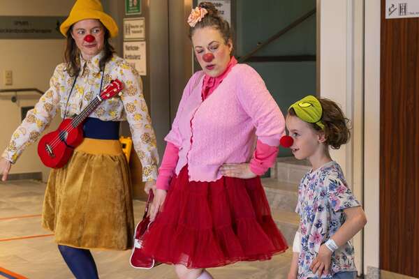 Zwei ROTE NASEN Clowns tanzen mit einem Kind auf einem Krankenhausflur, eine Clownin hält eine kleine rote Gitarre.