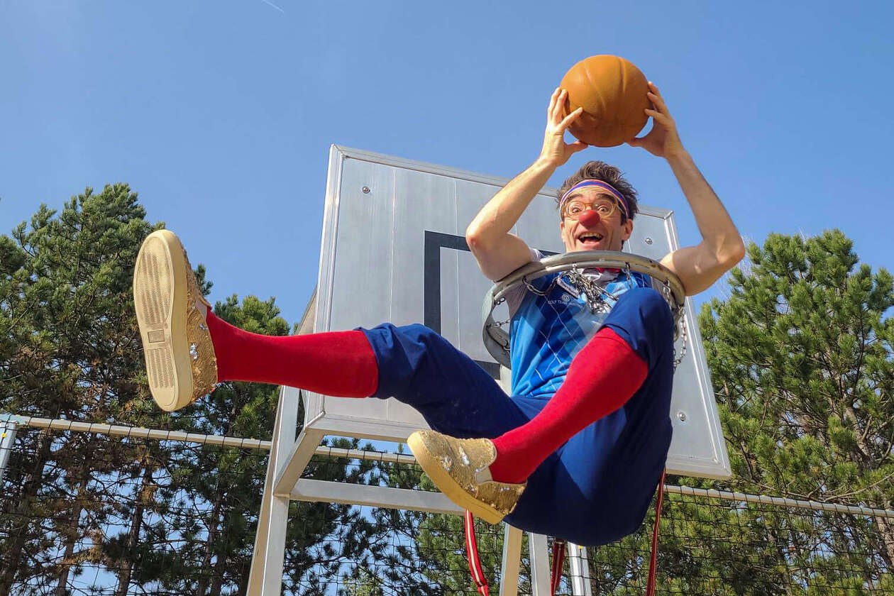 Clown Bernhart sitzt in einem Basketballkorb und hält einen Ball.