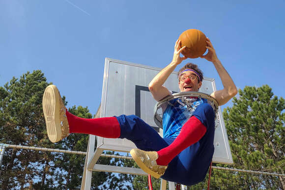 Clown Bernhart sitzt in einem Basketballkorb und hält einen Ball.