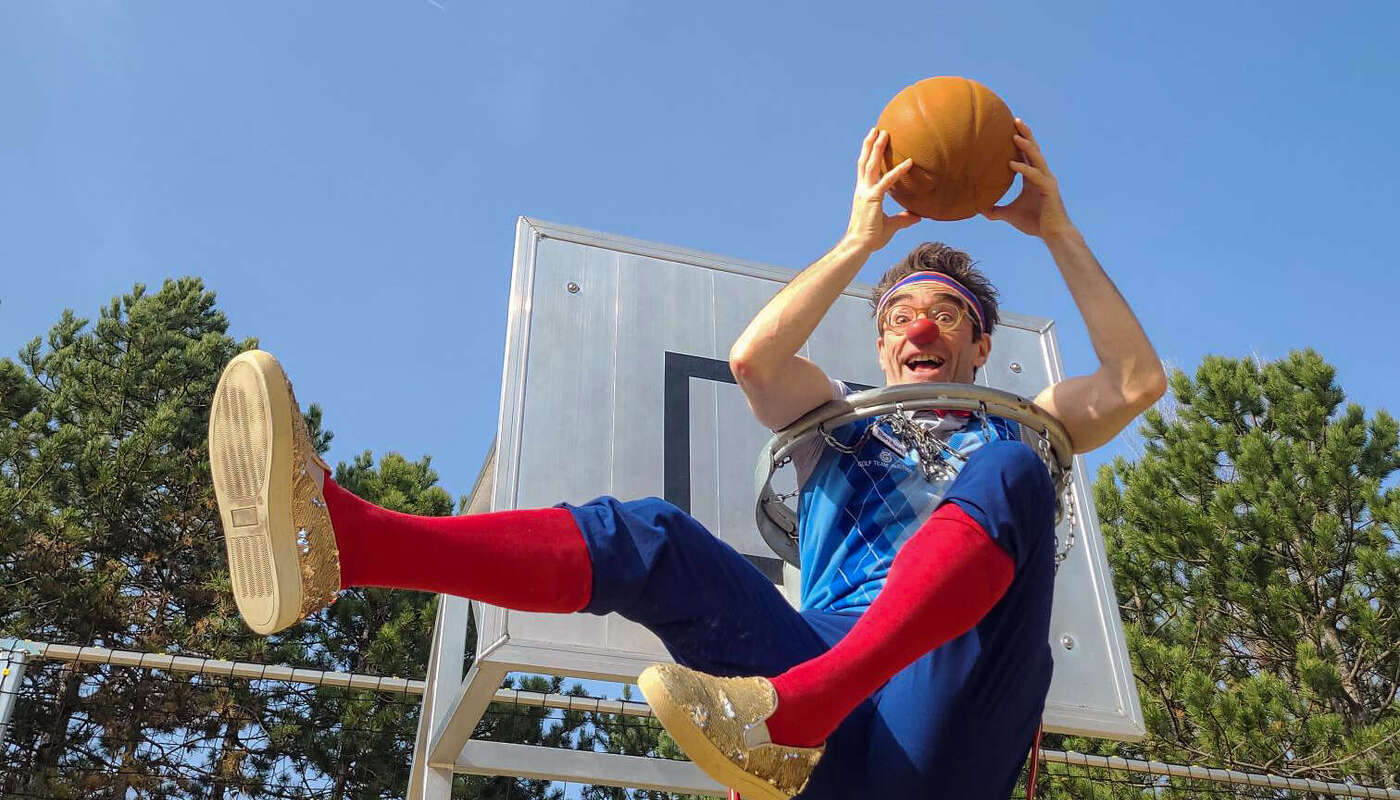 Clown Bernhart sitzt in einem Basketballkorb und hält einen Ball.