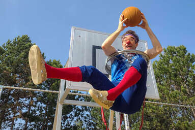 Clown Bernhart sitzt in einem Basketballkorb und hält einen Ball.