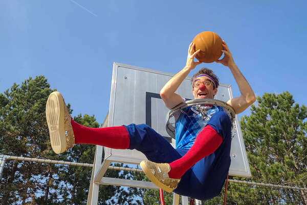 Clown Bernhart sitzt in einem Basketballkorb und hält einen Ball.