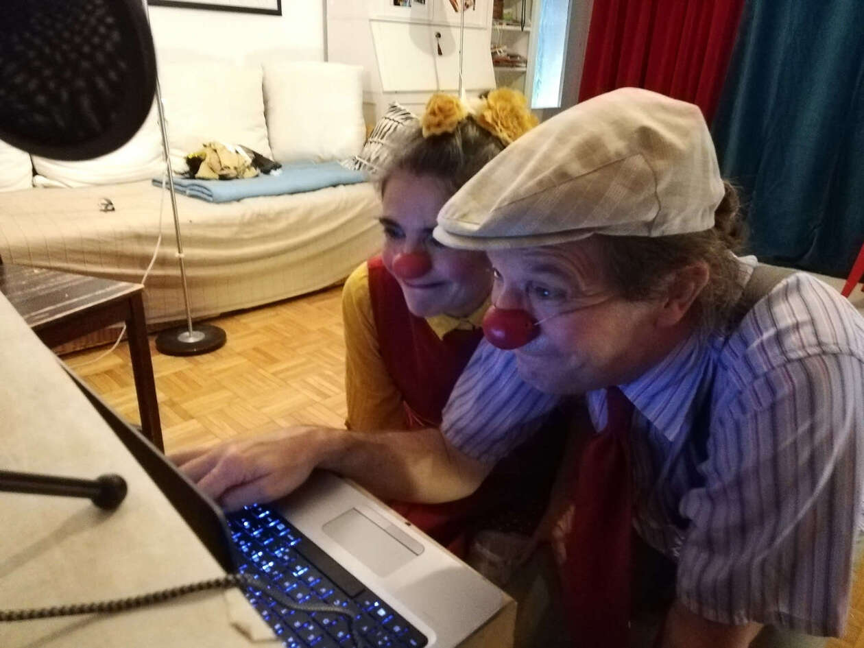 Clownin Rita sitzt mit Partner Guido vor dem Computer und lacht in die Kamera