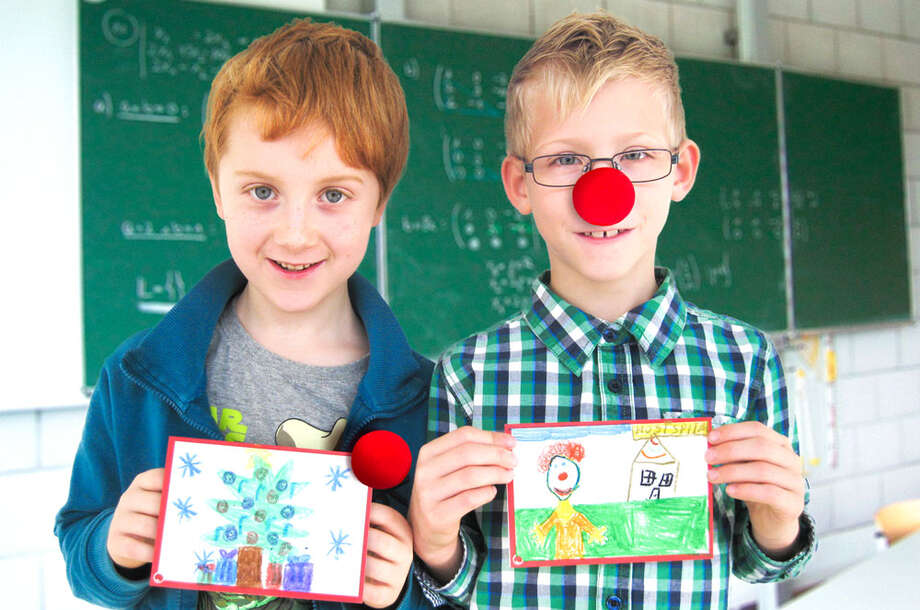 Zwei Schulkinder stehen vor einer Tafel und halten selbstgezeichnete Grußkarten, einer der Jungen trägt eine ROTE NASE