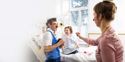 Ein Mädchen lacht bei einem Clownbesuch fröhlich im Krankenbett.