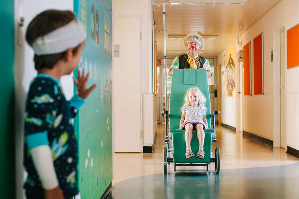 kleiner Junge steht mit Bandage am Kopf im Krankenhausgang und winkt einem kleinen Mädchen, das von einem Clown in einem Rollstuhl geschoben wird, zu.