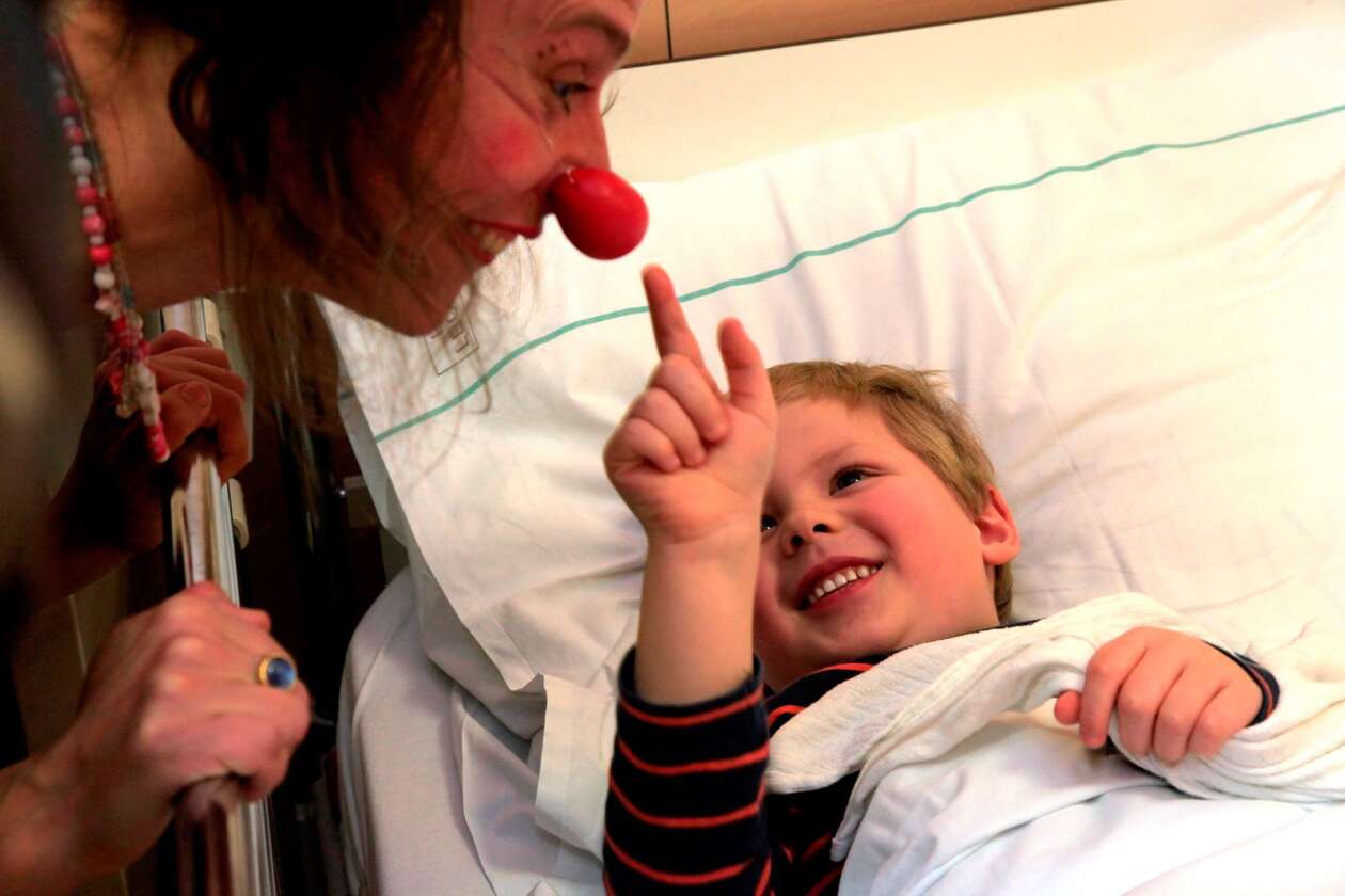 Clownin beugt sich zu einem kleinen Jungen im Spitalsbett, er greift ihr auf die Nase