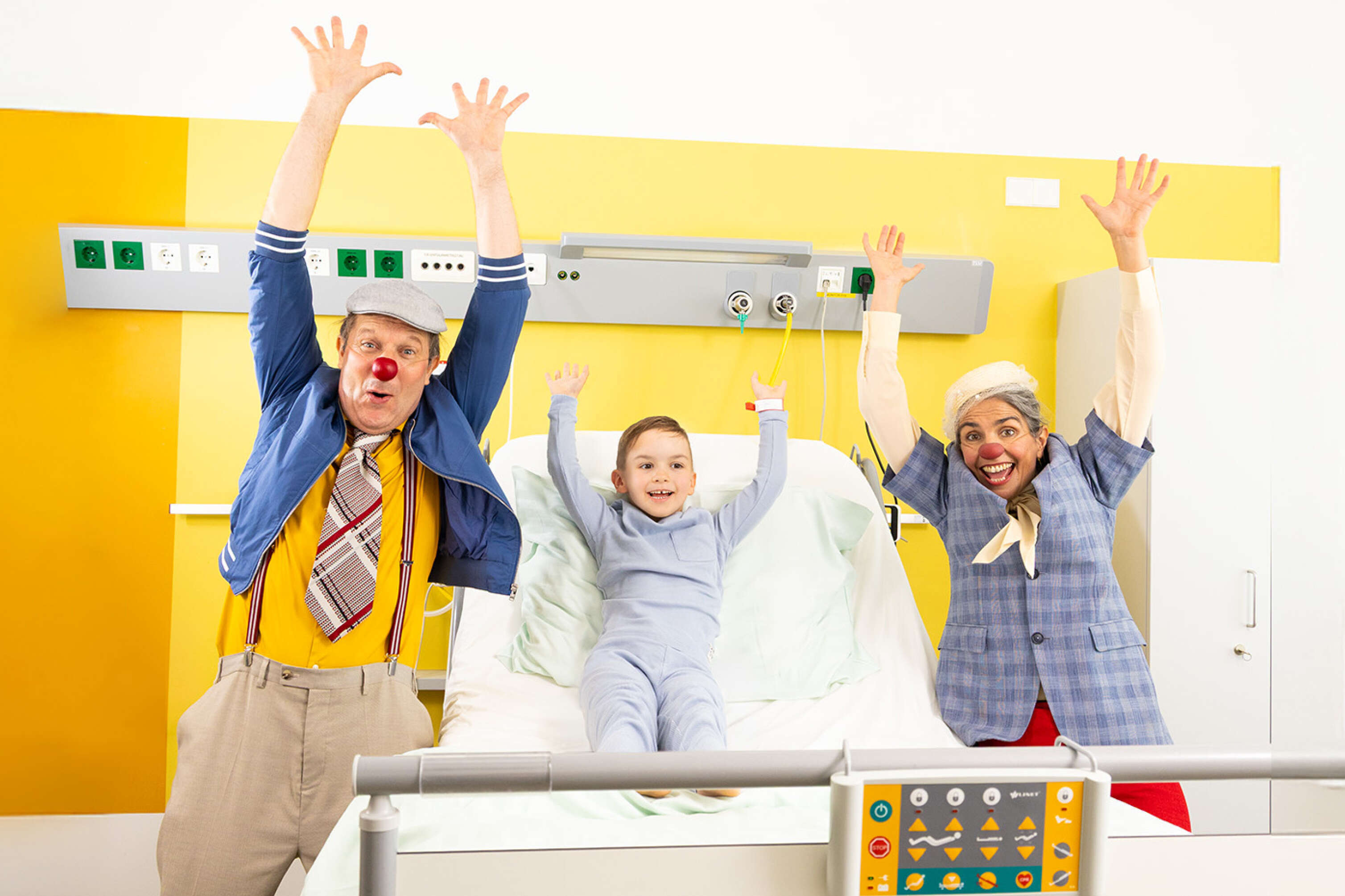 Zwei ROTE NASEN Clowns im Spital mit einem Kind und alle heben die Hände in die Luft.