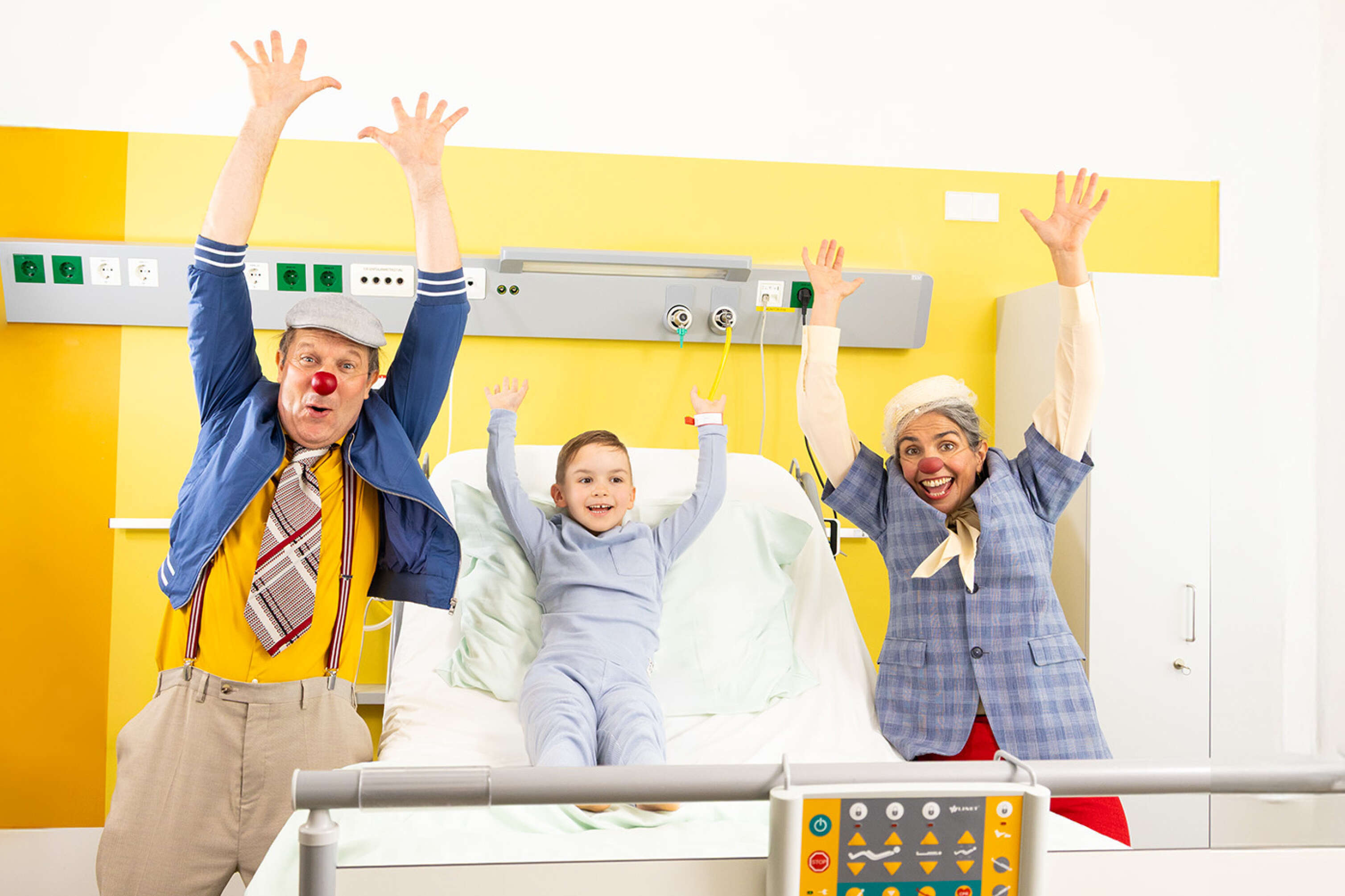 Zwei ROTE NASEN Clowns im Spital mit einem Kind und alle heben die Hände in die Luft.