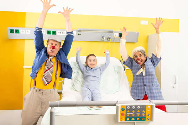 Zwei ROTE NASEN Clowns im Spital mit einem Kind und alle heben die Hände in die Luft.