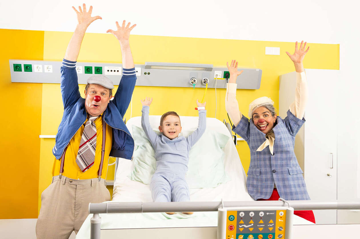 Zwei ROTE NASEN Clowns im Spital mit einem Kind und alle heben die Hände in die Luft.