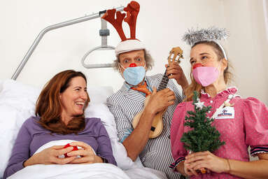 ROTE NASEN-Clowns Brösl mit Gitarre und Josefine mit Tannenbäumchen besuchen eine Frau an deren Krankenbett.