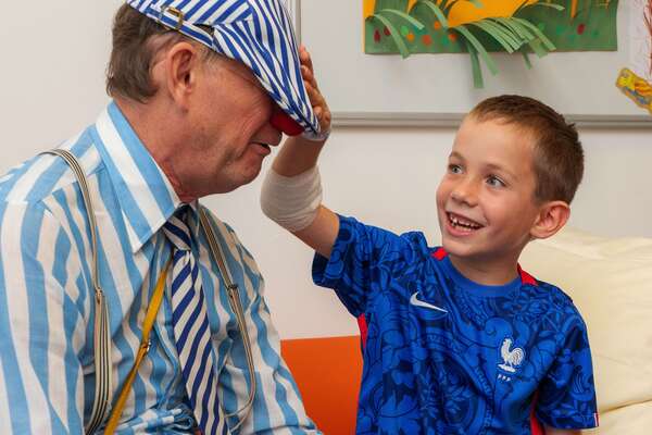 Ein ROTE NASEN Clown sitzt neben einem lachenden Jungen, der ihm spielerisch die gestreifte Mütze ins Gesicht zieht.