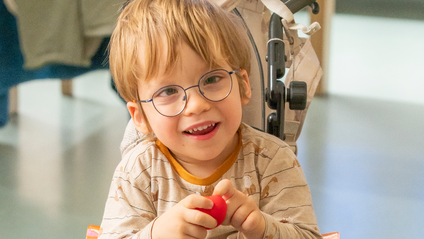 Junge im Rollstuhl lächelnd mit roter Nase in der Hand. 