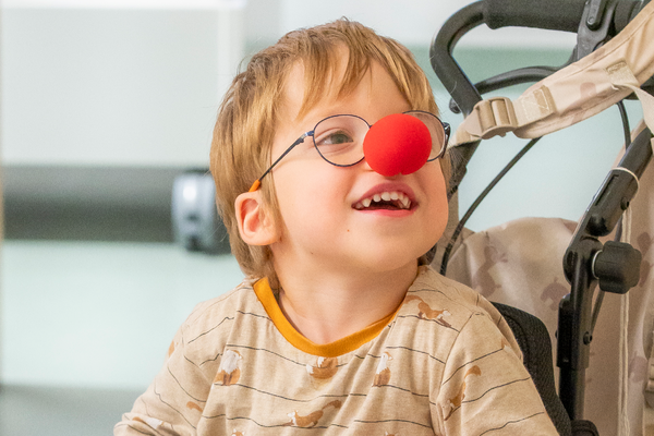 Junge im Rollstuhl lächelnd mit roter Clownnase auf seiner Nase.
