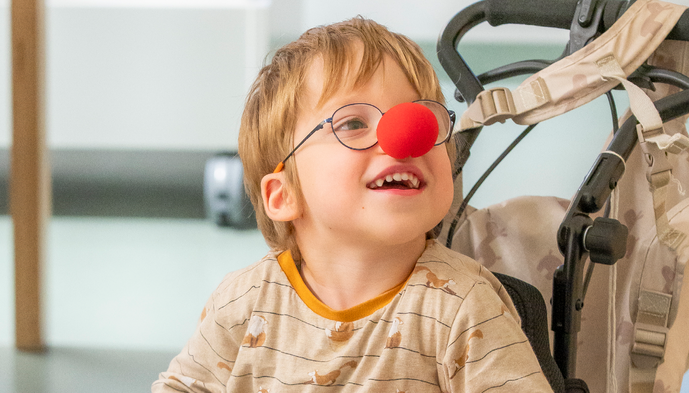 Junge im Rollstuhl lächelnd mit roter Clownnase auf seiner Nase.
