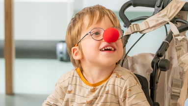 Junge im Rollstuhl lächelnd mit roter Clownnase auf seiner Nase.