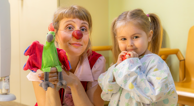 Ein kleines Mädchen mit einer roten Nase in der Hand neben Clownin Lilo und ihrer Handpuppe.