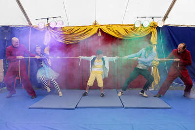 Ein Junge steht zwischen vier Clowns und hält an beiden Händen ein Seil, die Clowns ziehen von beiden Seiten an dem Seil