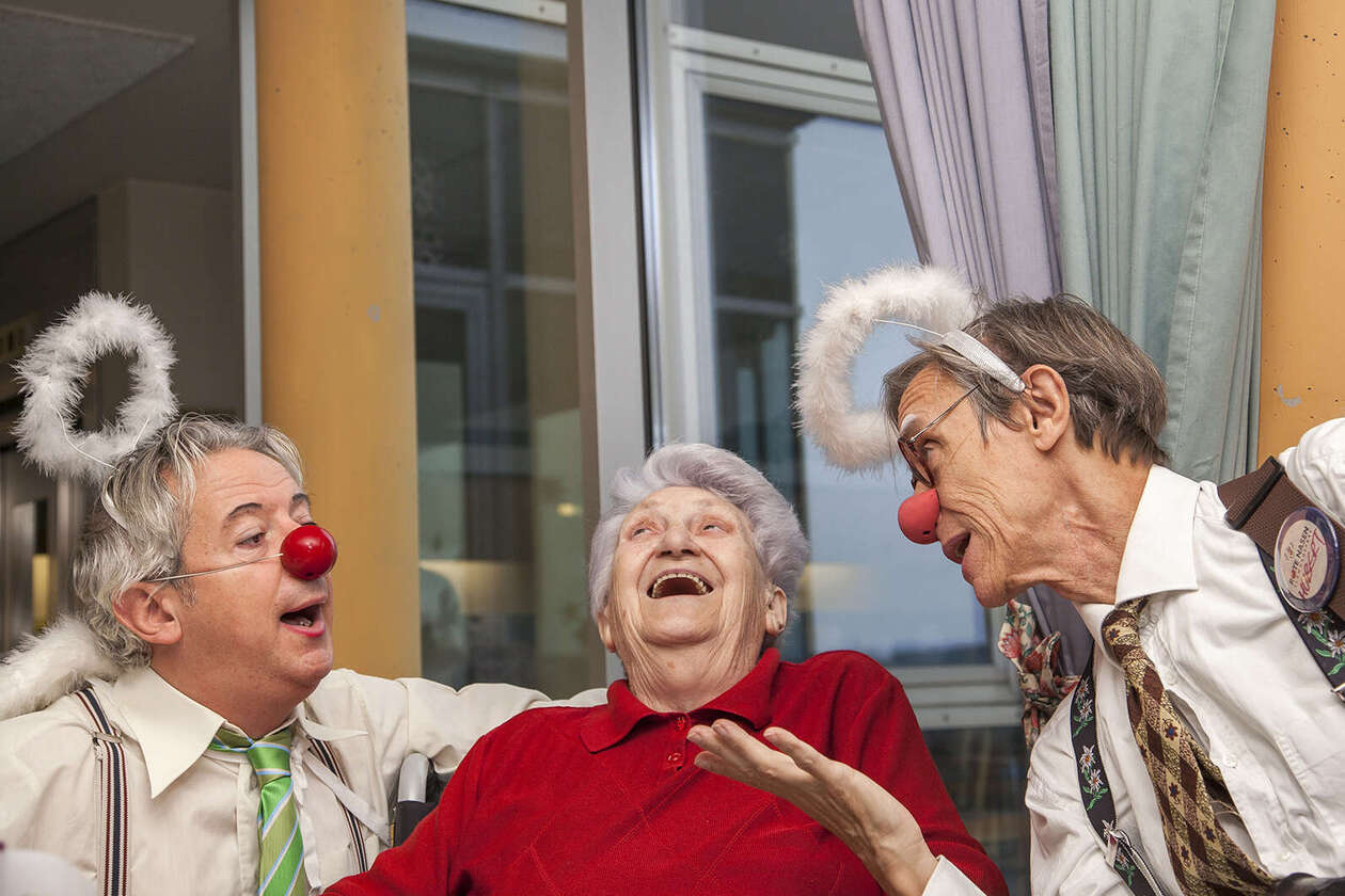 Seniorin sitzt zwischen zwei Clowns mit Engelsflügerln und Heiligenschein, sie singen gemeinsam und die Seniorin lacht herzlich