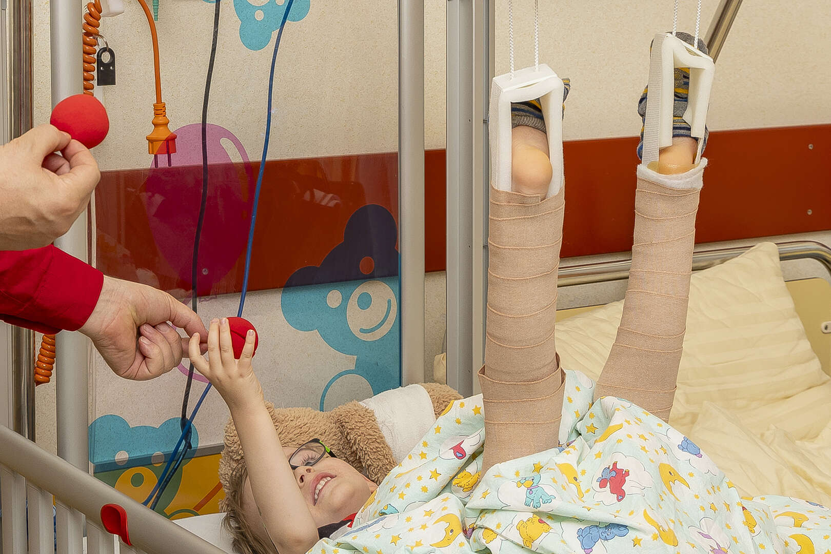 Kind im Krankenhausbett mit beidseitiger Beinschiene spielt lächelnd mit einer roten Schaumstoff Nase, die ihm von einer Person gereicht wird.