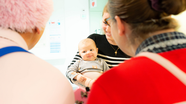 Zwei Clowns der ROTE NASEN stehen vor einer Mutter die ihr Baby im Arm hält.