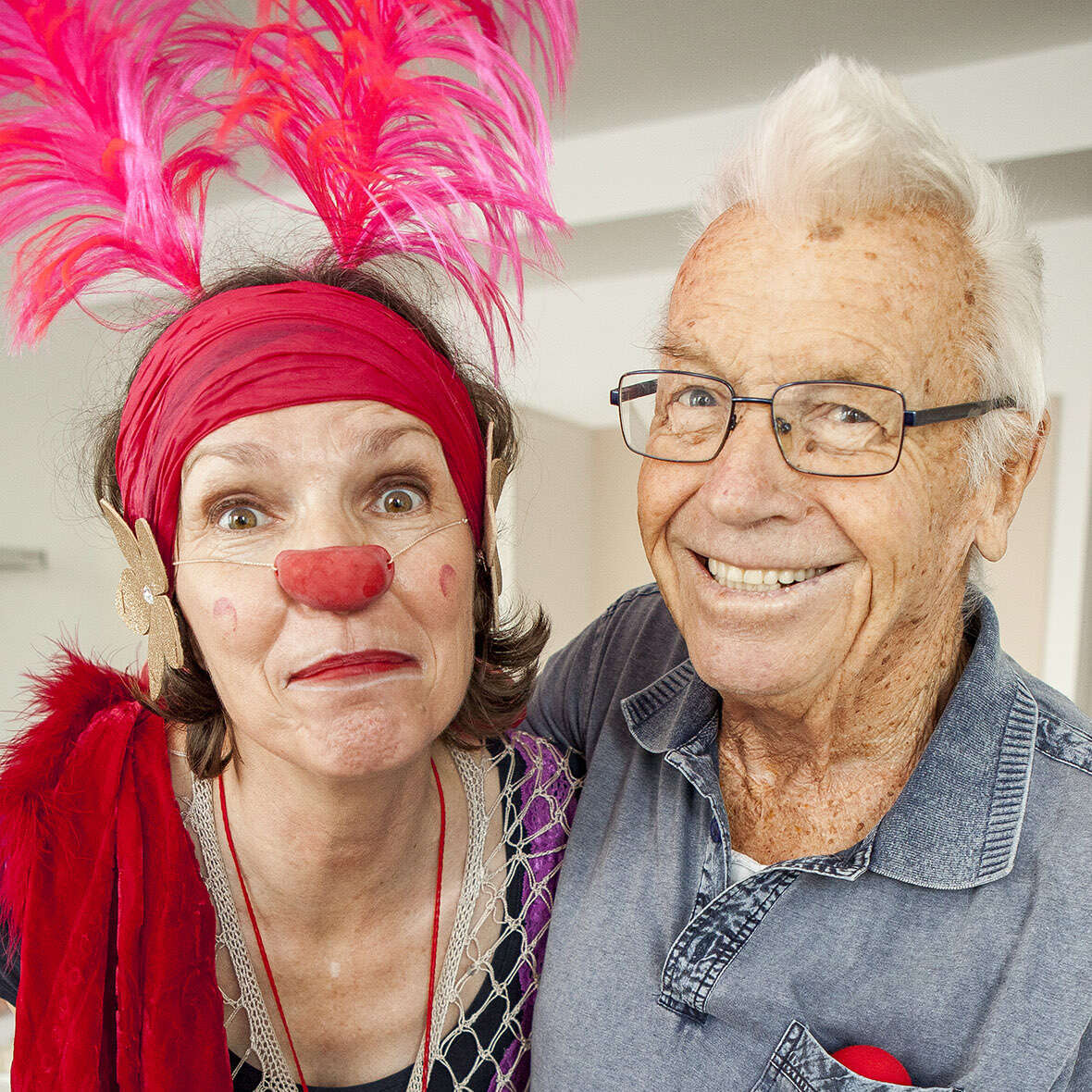 Eine Clownin mit Federn im Haar posiert gemeinsam mit einem Senior. Der Senior lacht herzlich in die Kamera.