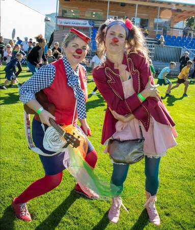 ROTE NASEN-Clownin Ecki mit Gitarre und Kollegin vor einer Tribüne während eines ROTE NASEN-Laufs.
