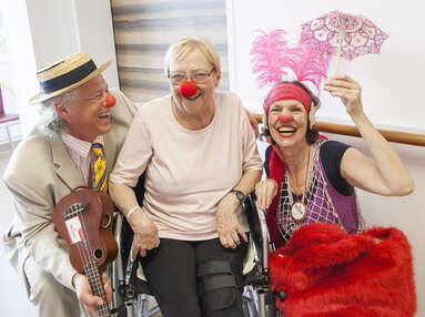 Eine Dame sitzt im Rollstuhl und trägt eine rote Schaumstoffnase. Links neben ihr sitzt ein Clown mit Ukulele in der Hand und lächelt sie herzhaft an. Rechts von ihr posiert eine Clownin mit Schirm und lächelt ebenfalls in die Kamera.