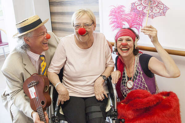Eine Dame sitzt im Rollstuhl und trägt eine rote Schaumstoffnase. Links neben ihr sitzt ein Clown mit Ukulele in der Hand und lächelt sie herzhaft an. Rechts von ihr posiert eine Clownin mit Schirm und lächelt ebenfalls in die Kamera.