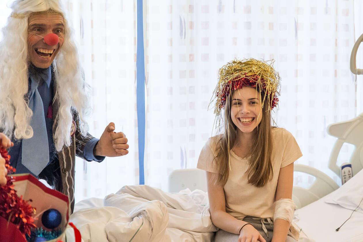 Clown mit weißer Langhaarperücke lacht herzlich, neben ihm sitzt ein Mädchen im Krankenhausbett und hat eine große Menge Lametta auf dem Kopf, sie lacht herzlich