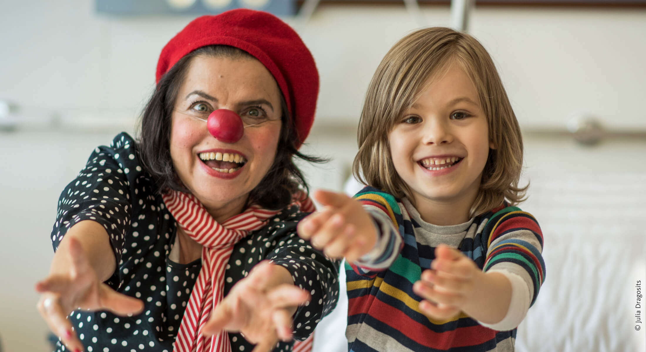 Ein Kind sitzt neben Clownin Olga und beide strecken mit fröhlichem Gesicht dem Betrachter die Hände entgegen.