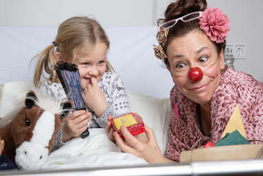 ROTE NASEN-Clownin Petra besucht ein Mädchen an ihrem Bett, dass einen Fächer in den Händen hält.