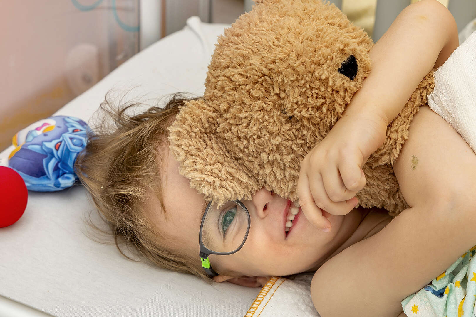 Kind im Bett liegend mit einem braunen Teddybär im Arm.