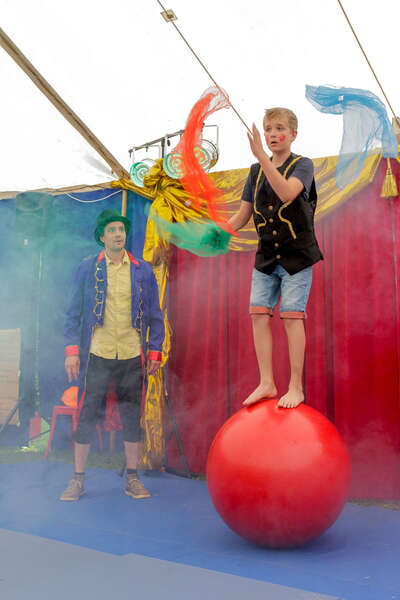 Ein Junge balanciert auf einem großen roten Ball und jongliert dabei mit bunten Tüchern