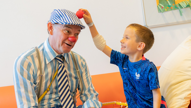 Ein kleiner Junge hält ROTE NASEN-Clown Herbert eine Schaumstoffnase auf dessen Kopf.