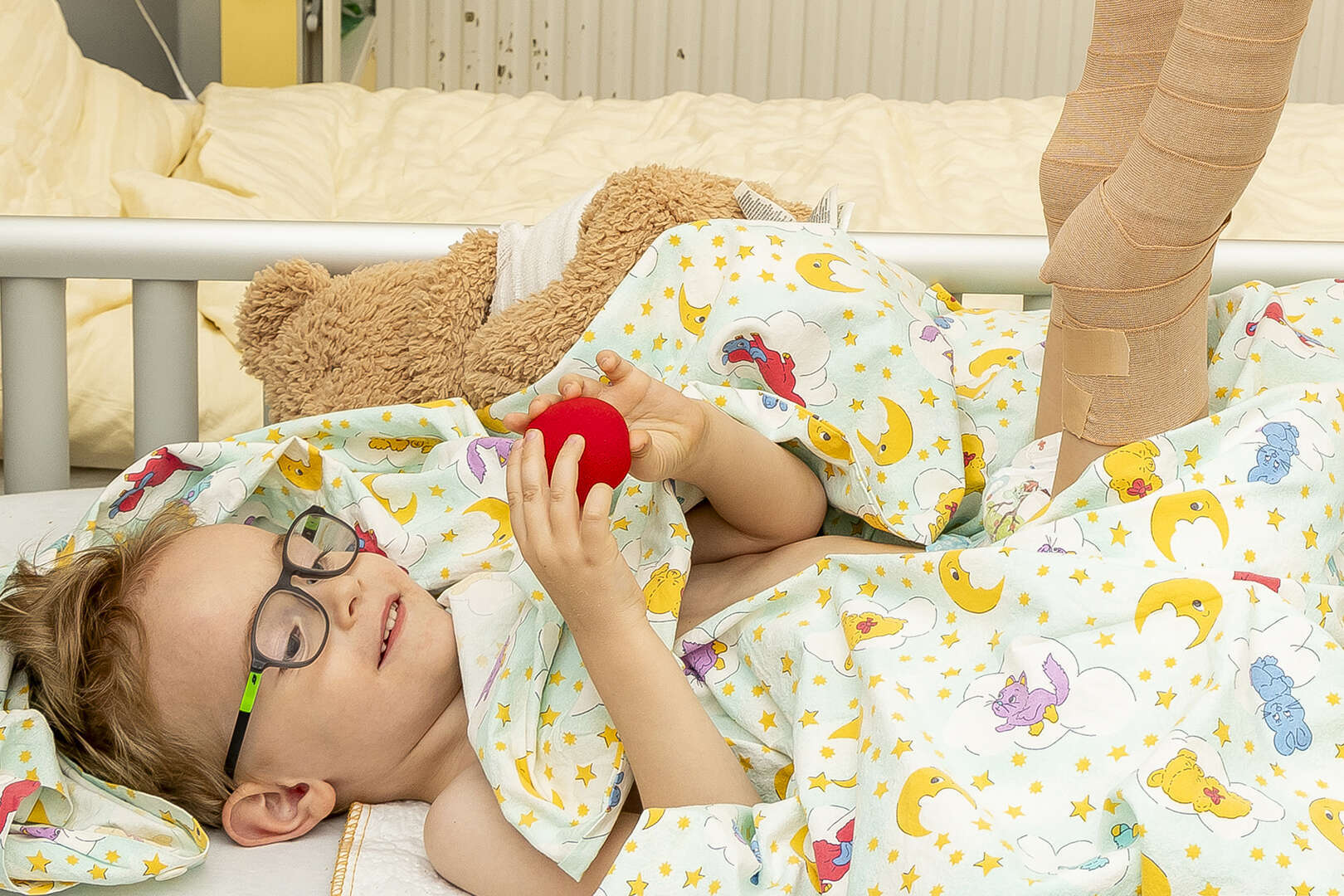 Kind im Krankenhausbett mit beidseitiger Beinschiene hält lächelnd eine rote Schaumstoffnase in den Händen, ein Teddybär liegt im Hintergrund.
