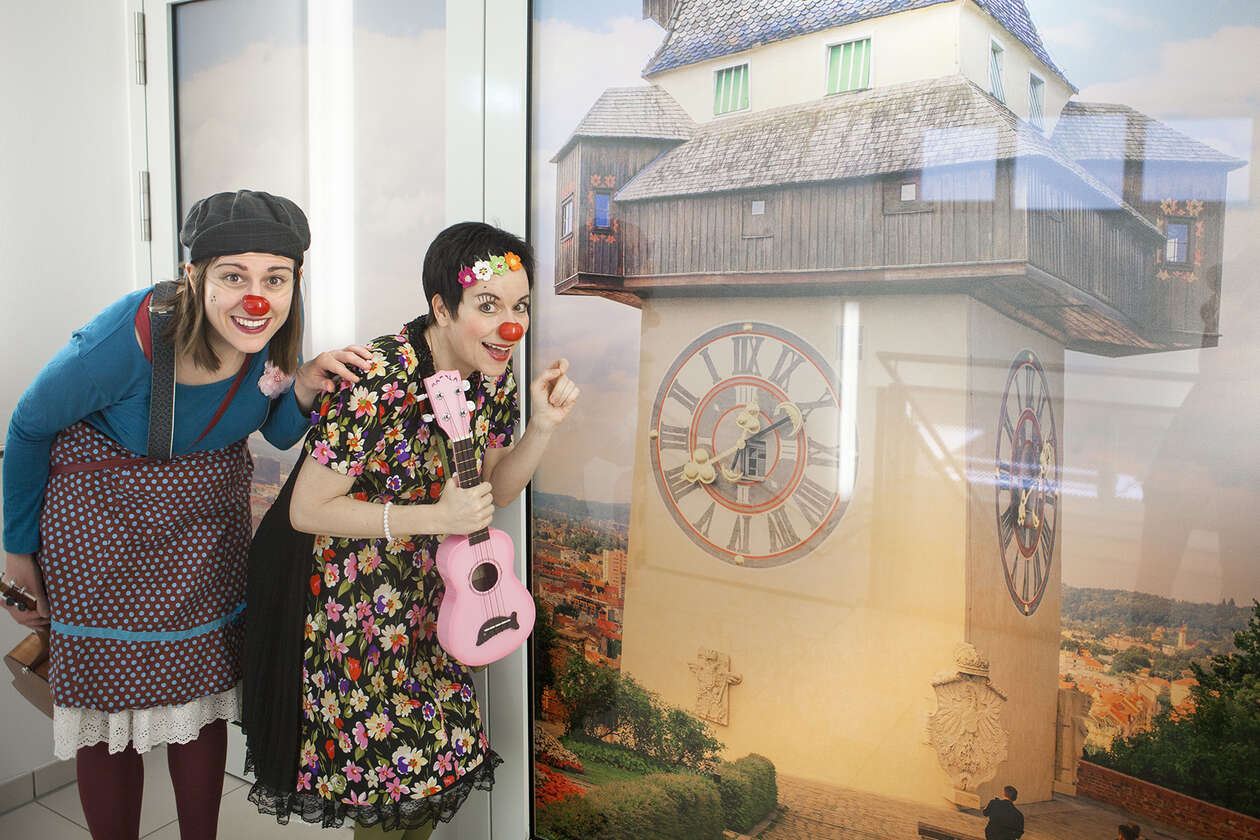 Zwei ROTE NASEN Clowninnen stehen hintereinander und leicht gebückt neben einem großen Foto des Grazer Uhrturms, eine Clownin hält eine rosa Ukulele in der Hand