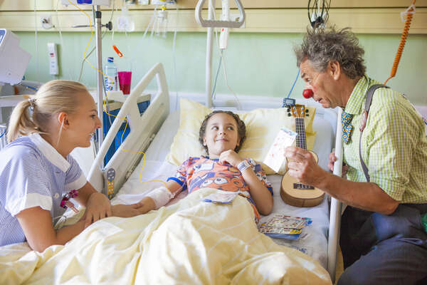 Clown und Krankenschwester sitzen bei Mädchen am Krankenbett