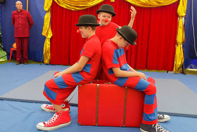 zwei Jungen mit Hüten und gestreiften Outfits sitzen auf einem roten Koffer, beide lassen die Köpfe hängen, ein dritter Junge sieht sie erwartungsvoll an und zuckt mit den Schultern
