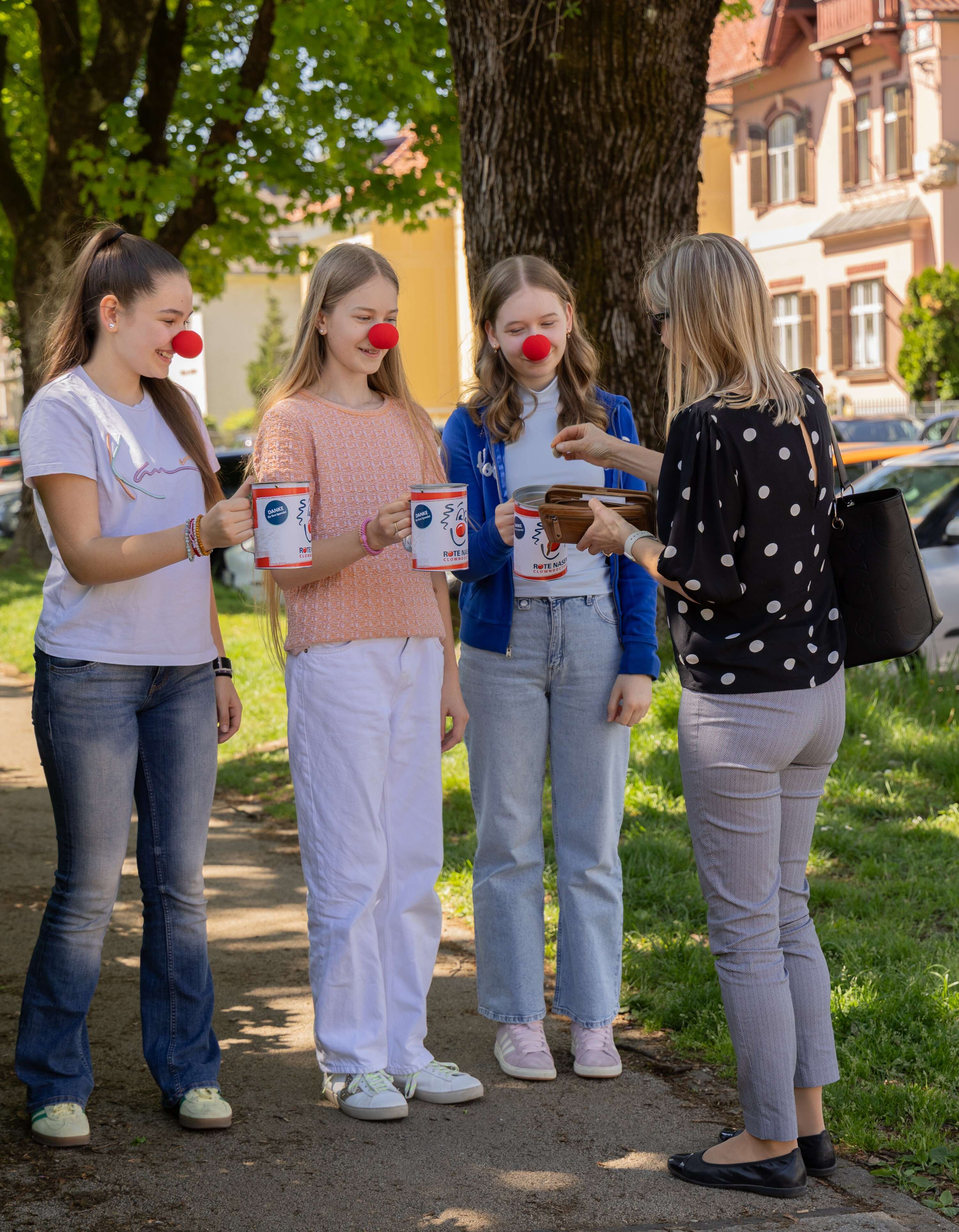 Drei junge Schülerinnen bei der ROTE NASEN Straßensammlung mit Spendendosen in der Hand und einer Spenderin.