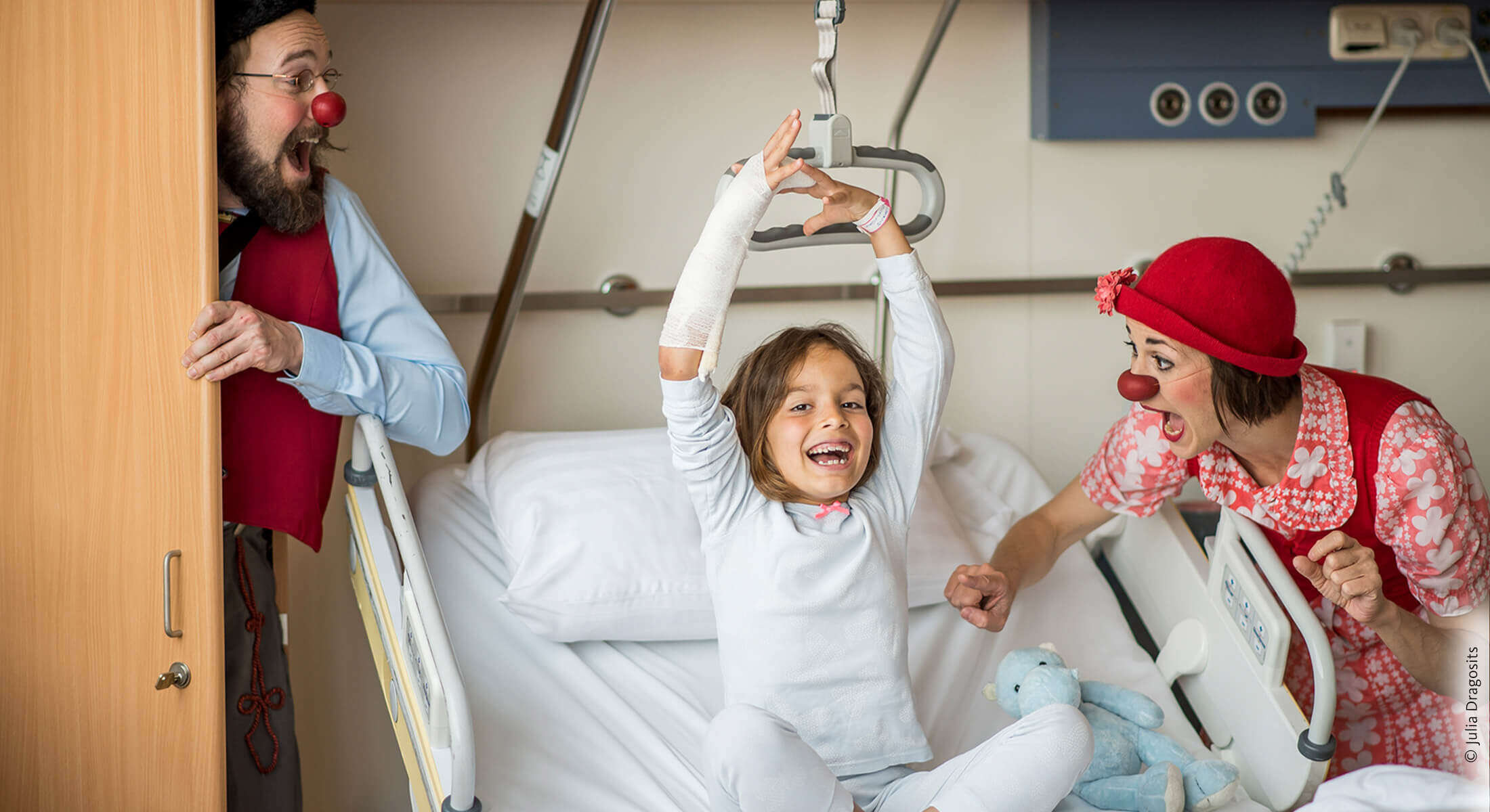 Kind sitzt im Spitalsbett und hebt fröhlich die Hände in die Luft während eine Clownin neben ihr sie zum Lachen bringt.