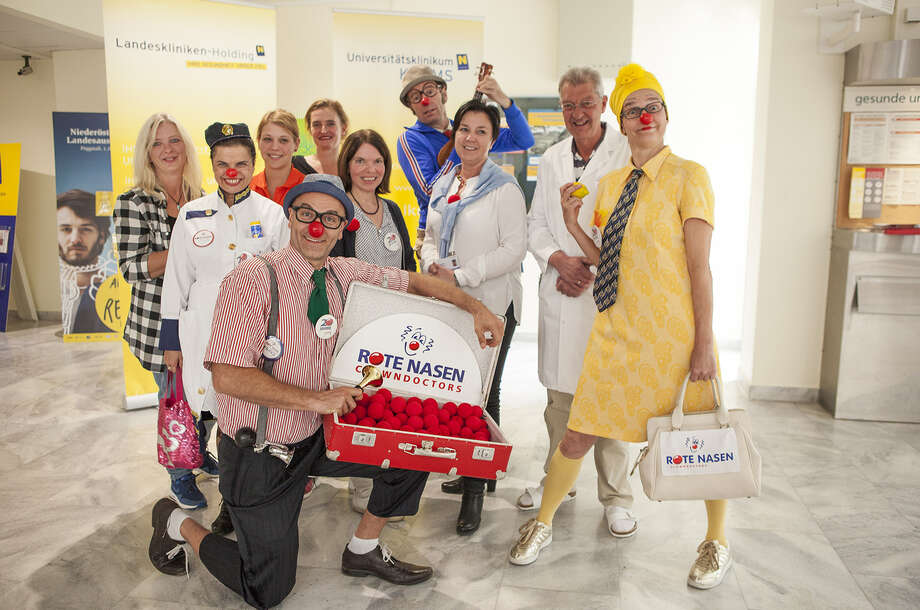 Gruppe aus Clowns, MitarbeiterInnen von ROTE NASEN und dem Krankenhaus Kremas posieren gemeinsam mit einem Koffer voll roten Schaumstoffnasen