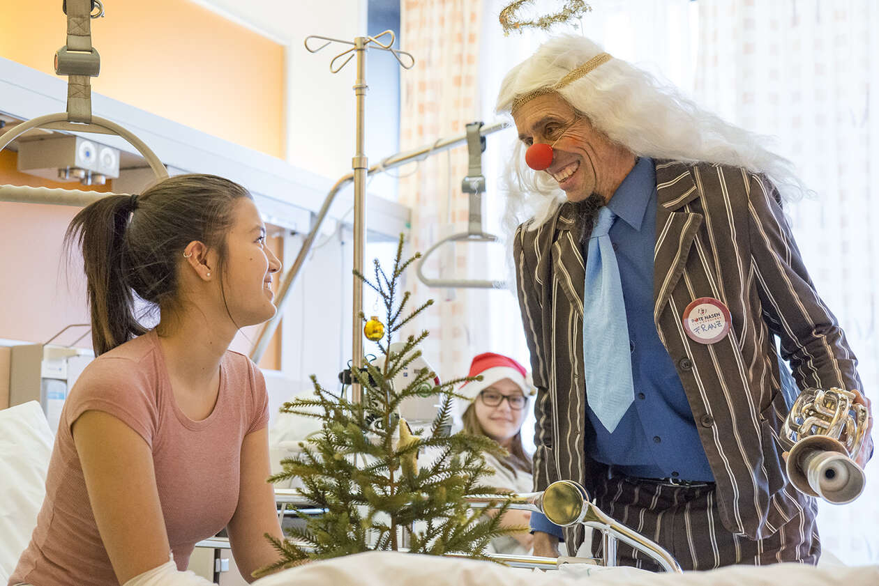 Clown mit weißer Langhaarperücke interagiert mit einem jungen Mädchen im Krankenhausbett, beide lachen und vor dem Mädchen steht ein kleiner Christbaum