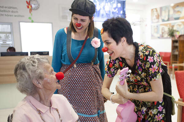 Seniorin sitzt im Sessel und trägt rote Schaumstoffnase, zwei Clowninnen stehen neben ihr und interagieren mit ihr.
