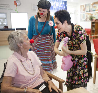 Seniorin sitzt im Sessel und trägt rote Schaumstoffnase, zwei Clowninnen stehen neben ihr und interagieren mit ihr.