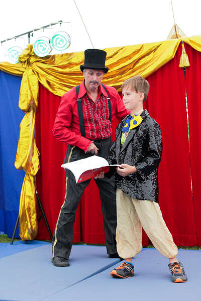 Ein Junge hält ein Buch in der Hand, neben ihm steht ein Clown in einem Zirkusdirektoroutfit