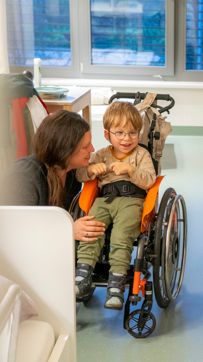Junge im Rollstuhl lächelt. Seine Mama sitzt daneben.