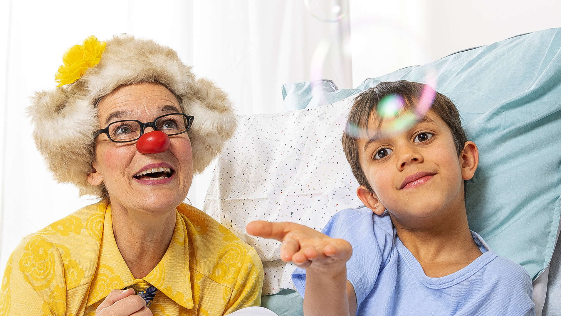 Eine ROTE NASEN Clownin mit roter Nase, gelbem Hemd und flauschiger Mütze mit Blume sitzt neben einem Jungen im Krankenhausbett. Beide lächeln und schauen einer Seifenblase nach.
